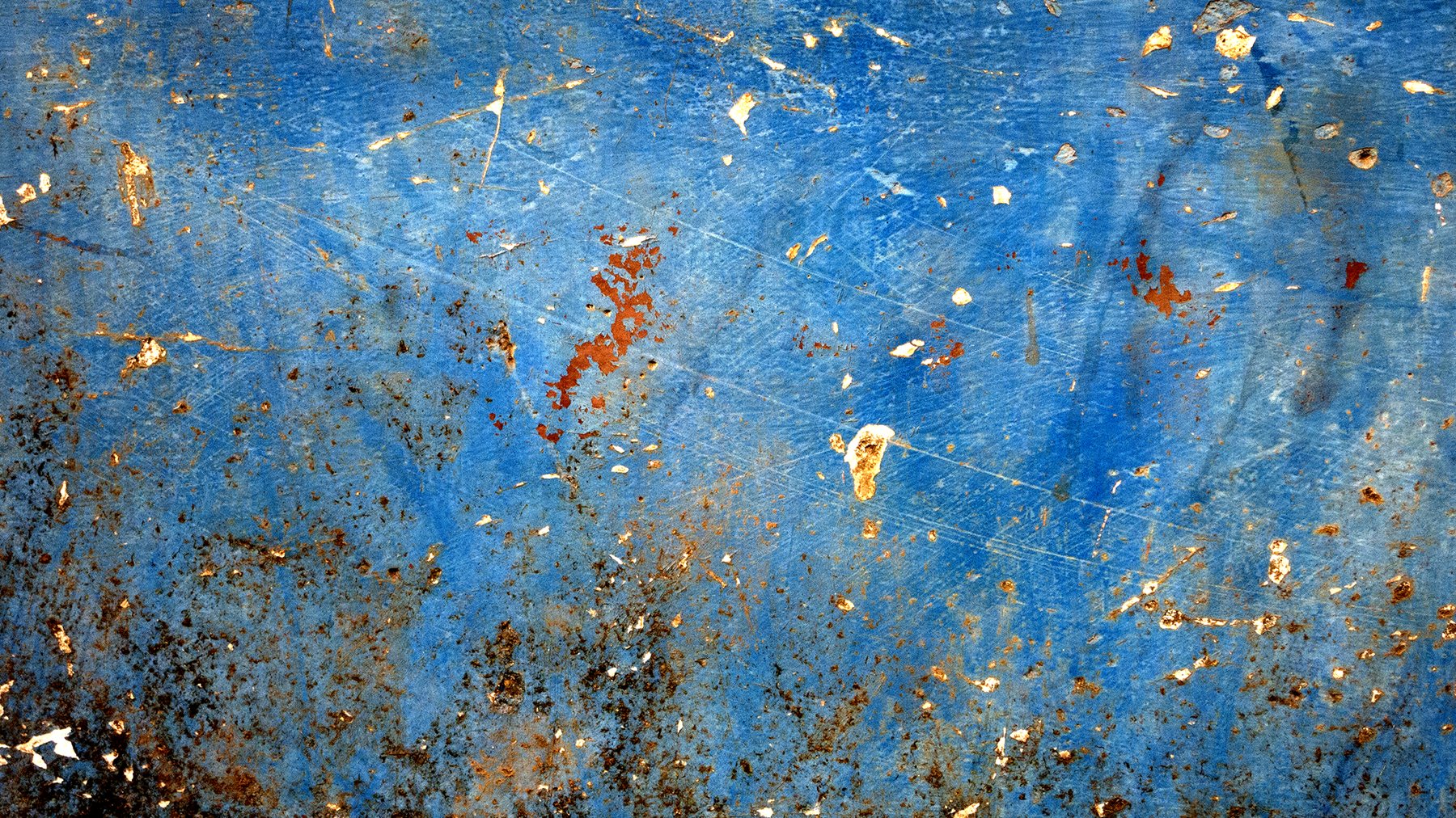 Blue painted boat hull, scratched and flecked with rust and pale chips, darkening to corrosion in one corner, photographed in Goa.